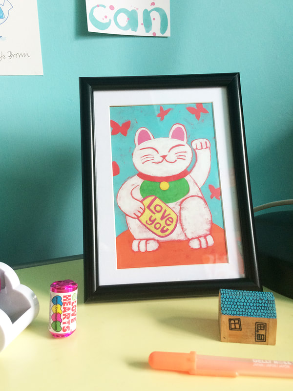 A framed illustrated postcard of a Lucky Cat waving by Jo Brown Illustrator on a messy desk.