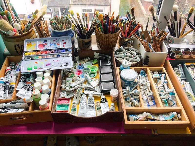 a desk with wooden trays filled with artists paint tubes, tin boxes of watercolours and various pots and cups filled with paintbrushes of different sizes