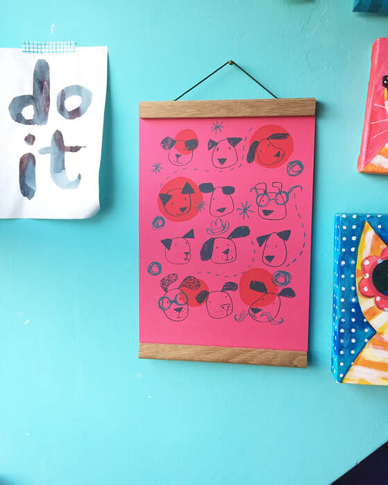 A pink print of dogs by Jo Brown, hanging on a turquoise wall with a handprinted piece of paper that says 'do it' taped to the wall next to it